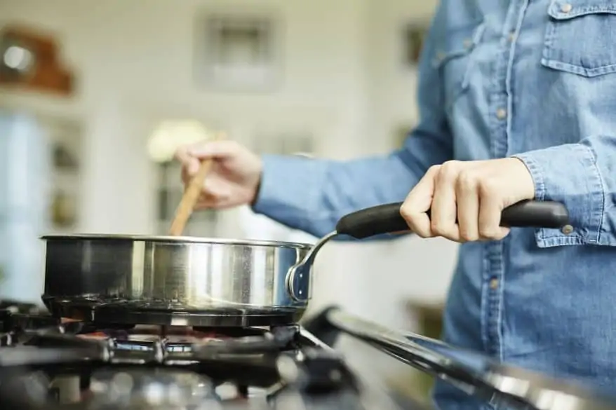 Midsection image of woman cooking food in frying pan. Utensil is placed on gas stove. Female is stirring dish in cooking pan. She is preparing food in domestic kitchen.