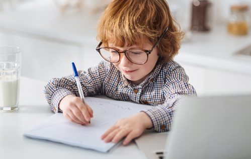 getting ready for school untypical school year. Persistent lively ginger kid carefully writing down his thoughts while wearing big glasses and sitting at white table