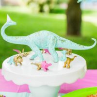 dinosaur figure wearing party hat on cake stand surrounded by small Dinos