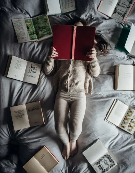 A girl reading on a bed with books all around her.
