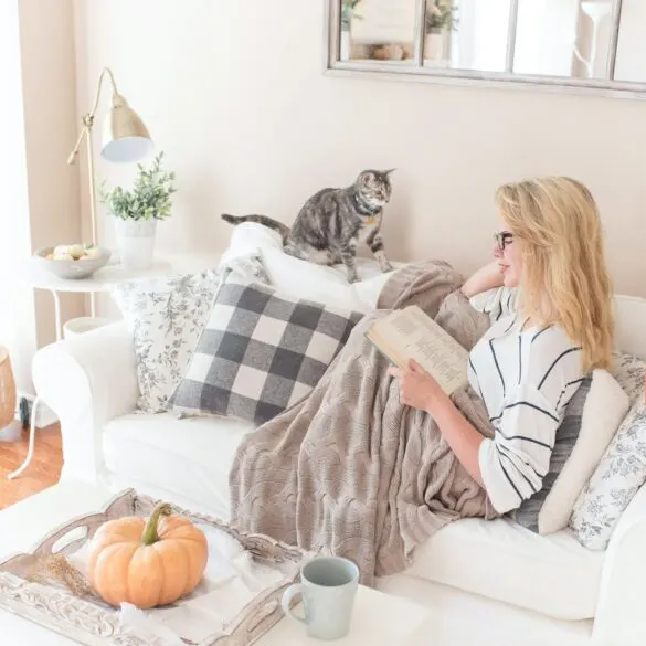 A lady on a couch with a blanket and a cat reading.