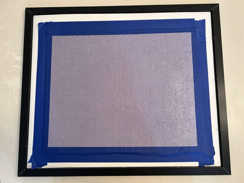 A puzzle face down in a frame with blue painters tape around the edges to secure it.