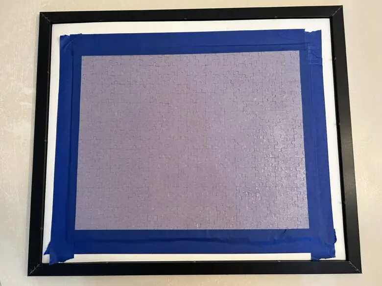 A puzzle face down in a frame with blue painters tape around the edges to secure it.